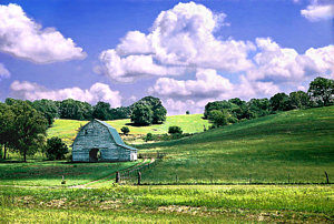 Image of a countryside in an unnamed Missouri town. THe grass is green and there is a wooden barn and bright white clouds.